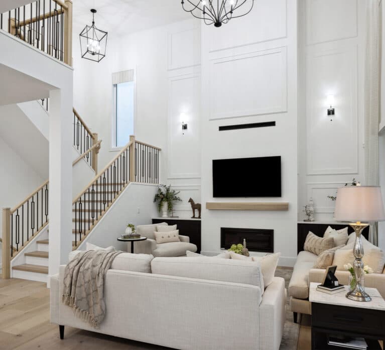 Brookswood Estates – A great room with a two-story ceiling, wood-and-iron staircase, wall-mounted TV above a fireplace, and a large sectional sofa.