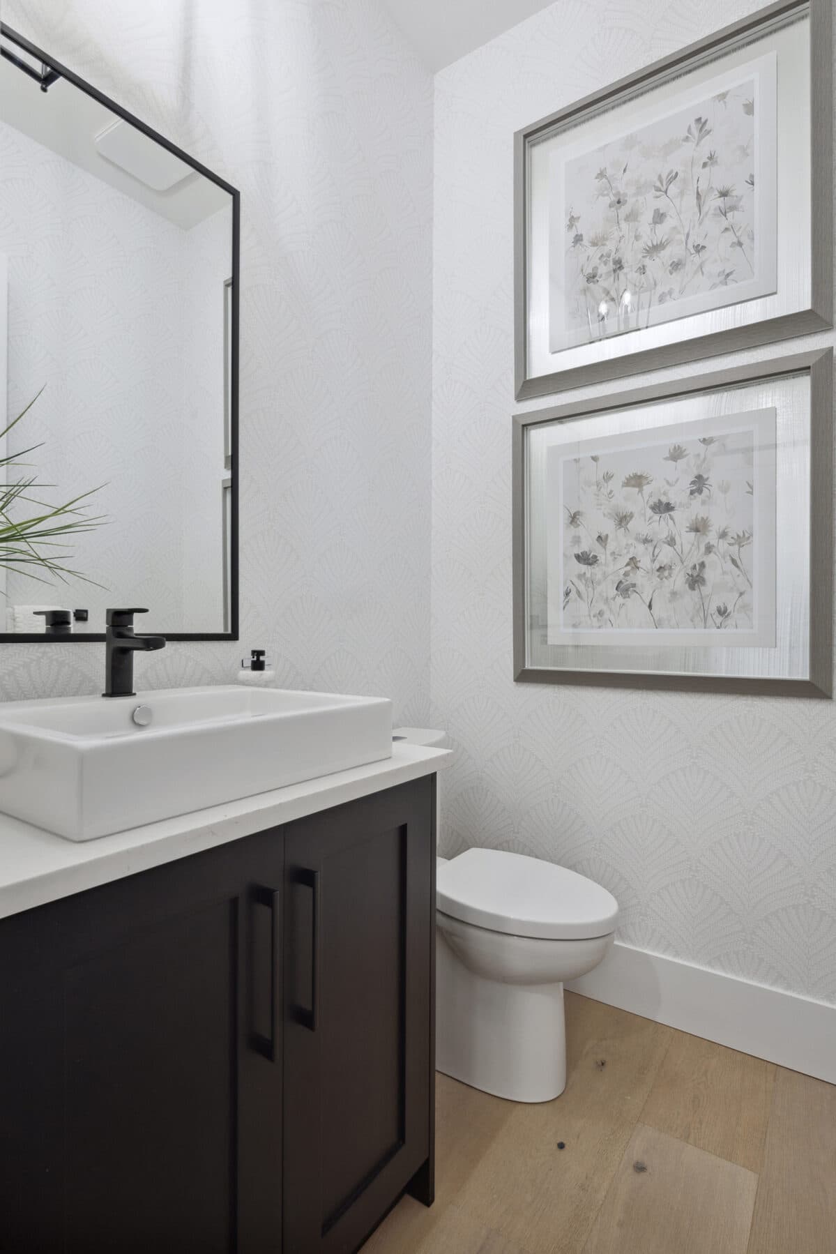 Brookswood Estates – A powder room with a dark wood vanity, white vessel sink, black fixtures, patterned wallpaper, and two framed floral prints above the toilet.