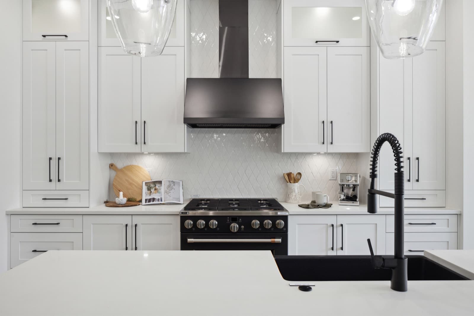 Brookswood Estates – A close-up kitchen view of white shaker cabinets, a black range hood, tile backsplash, and a black island sink with faucet in the foreground.