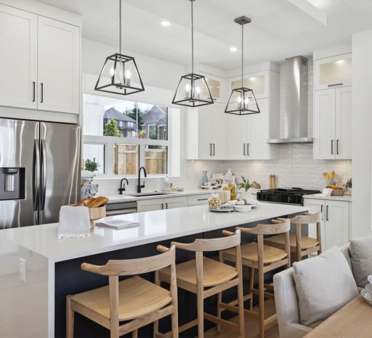 Brookswood Estates – A white kitchen with a large island, wood bar stools, pendant lighting, and a view through a window to a neighboring house, with a sofa visible in the foreground.