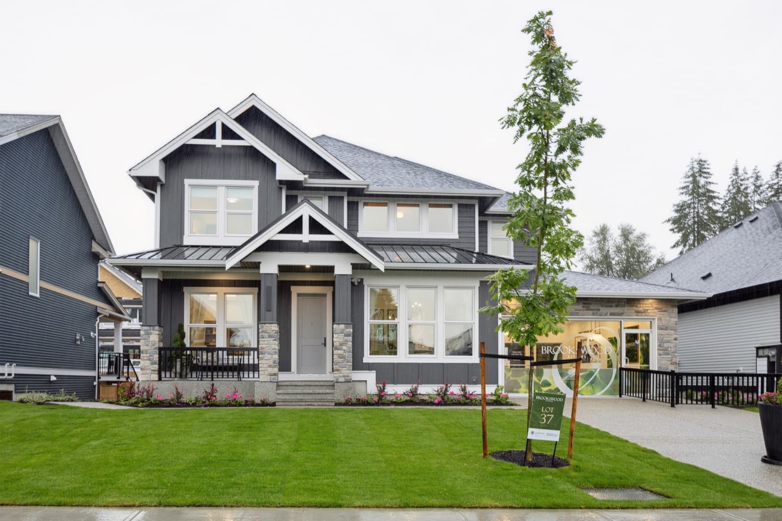 Brookswood Estates – A two-story craftsman-style show home exterior in dark grey siding with white trim, stone accents, a covered front porch, and a landscaped front lawn with a Brookswood lot sign.