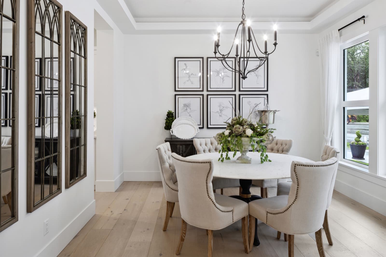 Brookswood Estates – A dining room with a round marble-top table, upholstered nailhead chairs, an iron chandelier, and a gallery wall of framed botanical sketches.