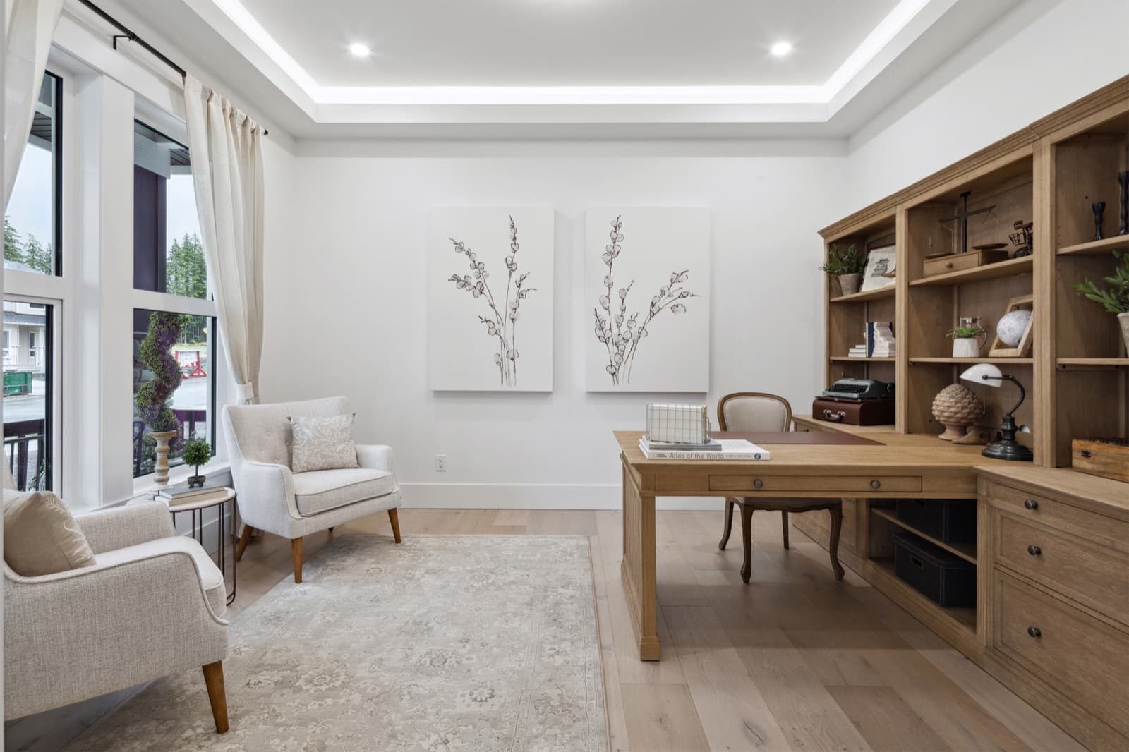 Brookswood Estates – A home office with a wood desk and chair, built-in wood shelving with decorative objects, two armchairs, and framed botanical art on the wall.