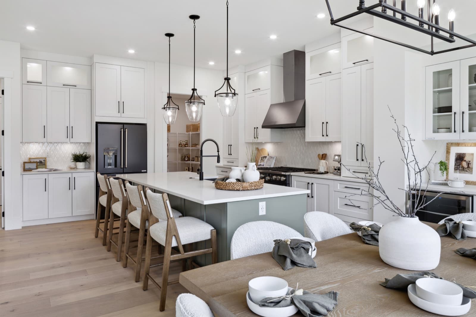 Brookswood Estates – An open kitchen with white cabinetry, a dark green island with wood bar stools, black pendant lights, and a black French-door refrigerator, opening onto a dining area.
