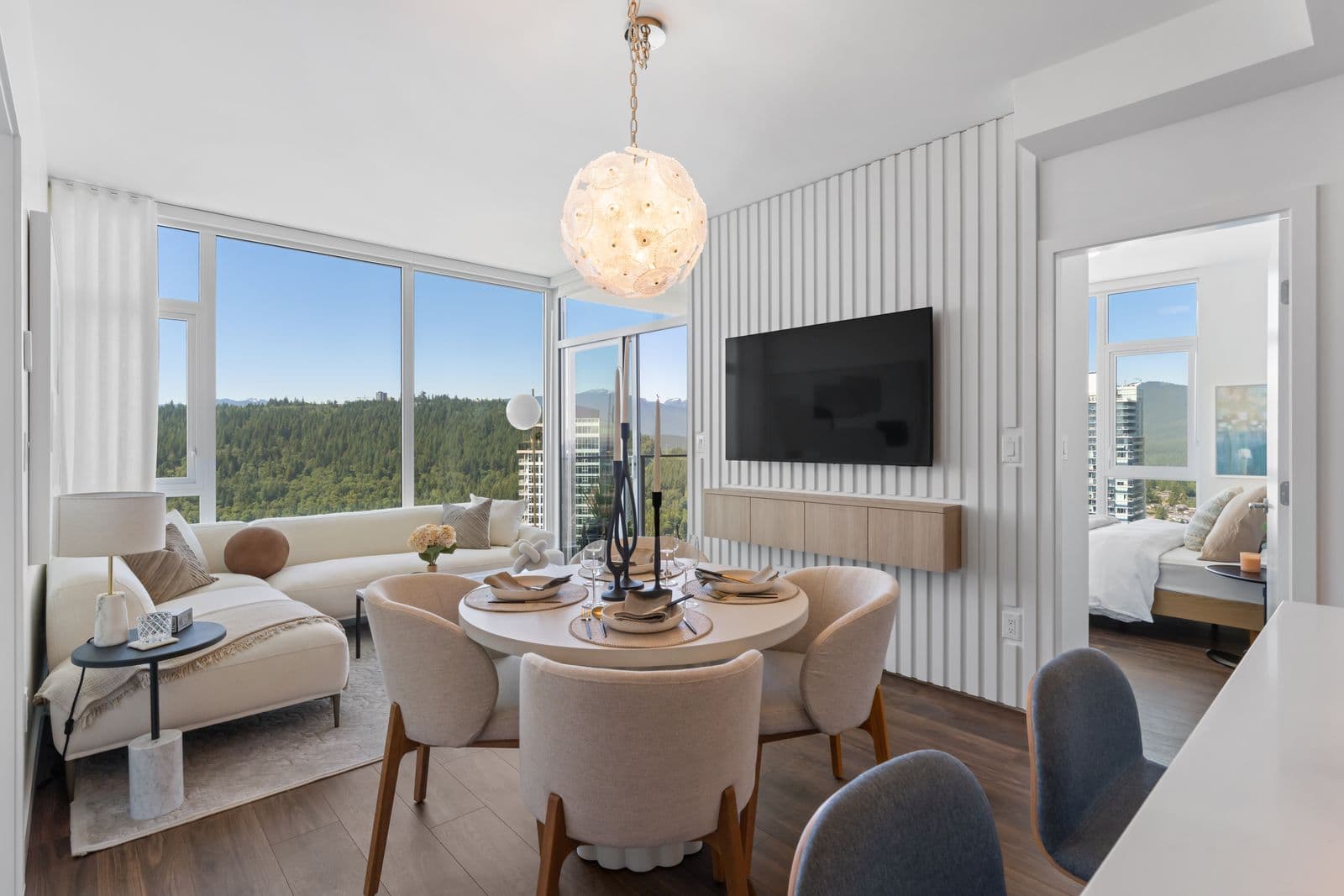 Highpoint Condos – Living and dining area with a round table set for a meal, a white sectional chaise, and a wall-mounted TV, with a bedroom visible through a doorway and forested mountain views through the windows.