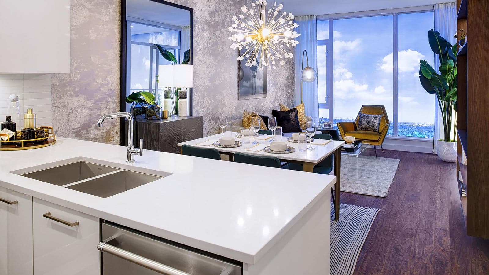 Highpoint Condos – Kitchen, dining, and living area with a white waterfall island, gold starburst chandelier over a set dining table, and a mustard accent chair by floor-to-ceiling windows overlooking the city.
