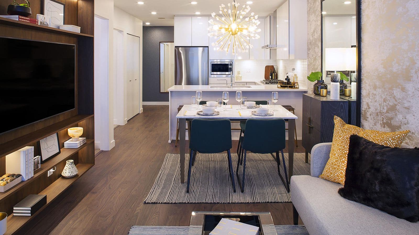 Highpoint Condos – Open-concept unit with a dining table set for four beneath a gold starburst chandelier, a white kitchen with stainless refrigerator, and a media wall with a mounted TV.