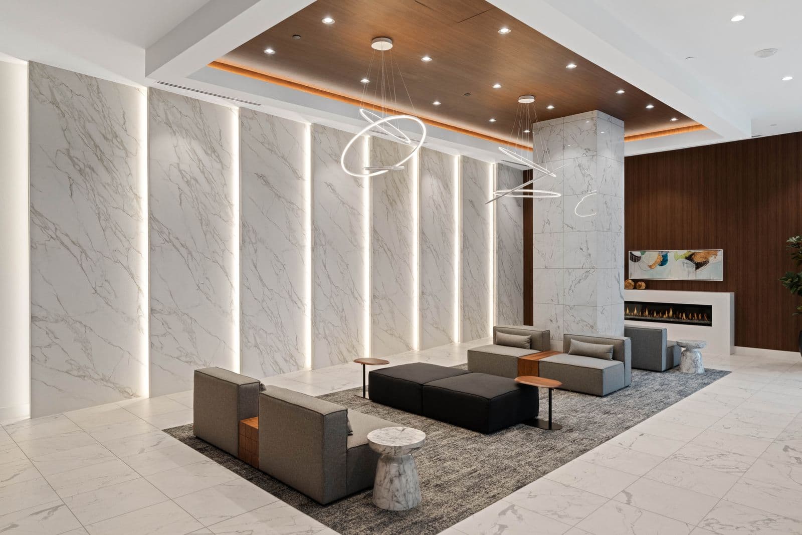 Highpoint Condos – Lobby lounge with marble feature wall, circular pendant lights, grey modular sofas around a black ottoman, and a gas fireplace along a wood-panelled wall.