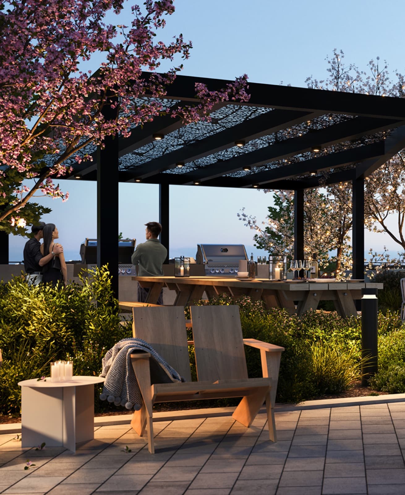 Encore – An upscale rooftop terrace at dusk featuring a black metal pergola with integrated lighting over a long wooden dining table and built-in BBQ stations. Lush landscaping, blooming cherry blossom trees, and cozy wooden lounge chairs with a throw blanket create an inviting, warm atmosphere for residents.
