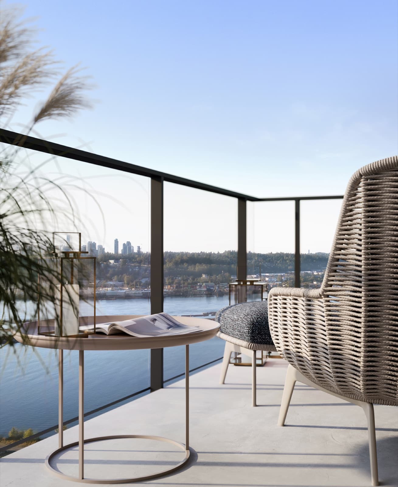 Encore – A modern high-rise balcony features a woven lounge chair, a matching footrest, and a small round side table with a book and lantern. Through the glass railing, a scenic daytime view unfolds over a wide river, forested hills, and a distant city skyline under a clear blue sky.