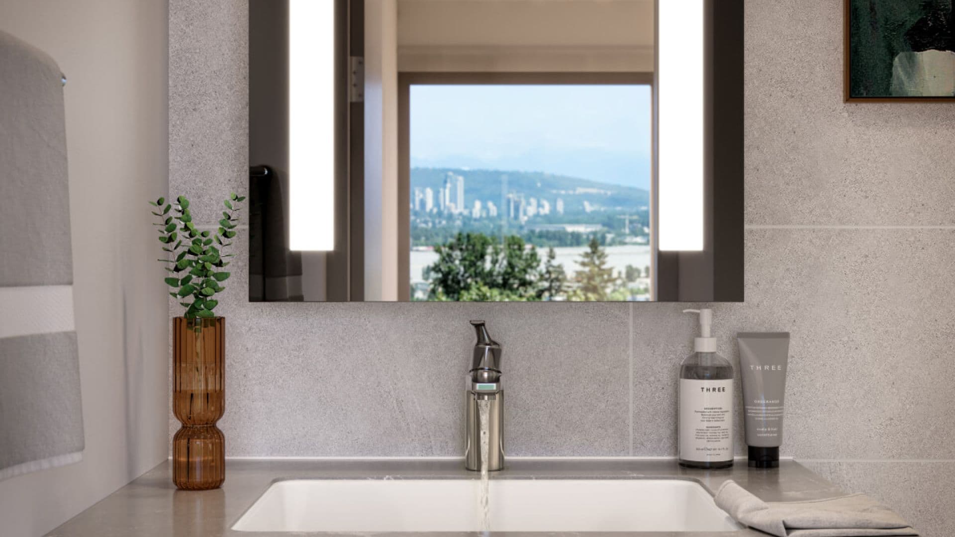 BridgeCity – A modern bathroom vanity features a running brushed-metal faucet, light gray stone backsplash, and an illuminated mirror reflecting an outdoor city skyline with distant mountains. A ribbed amber glass vase with eucalyptus and high-end toiletries frame the undermount white sink.