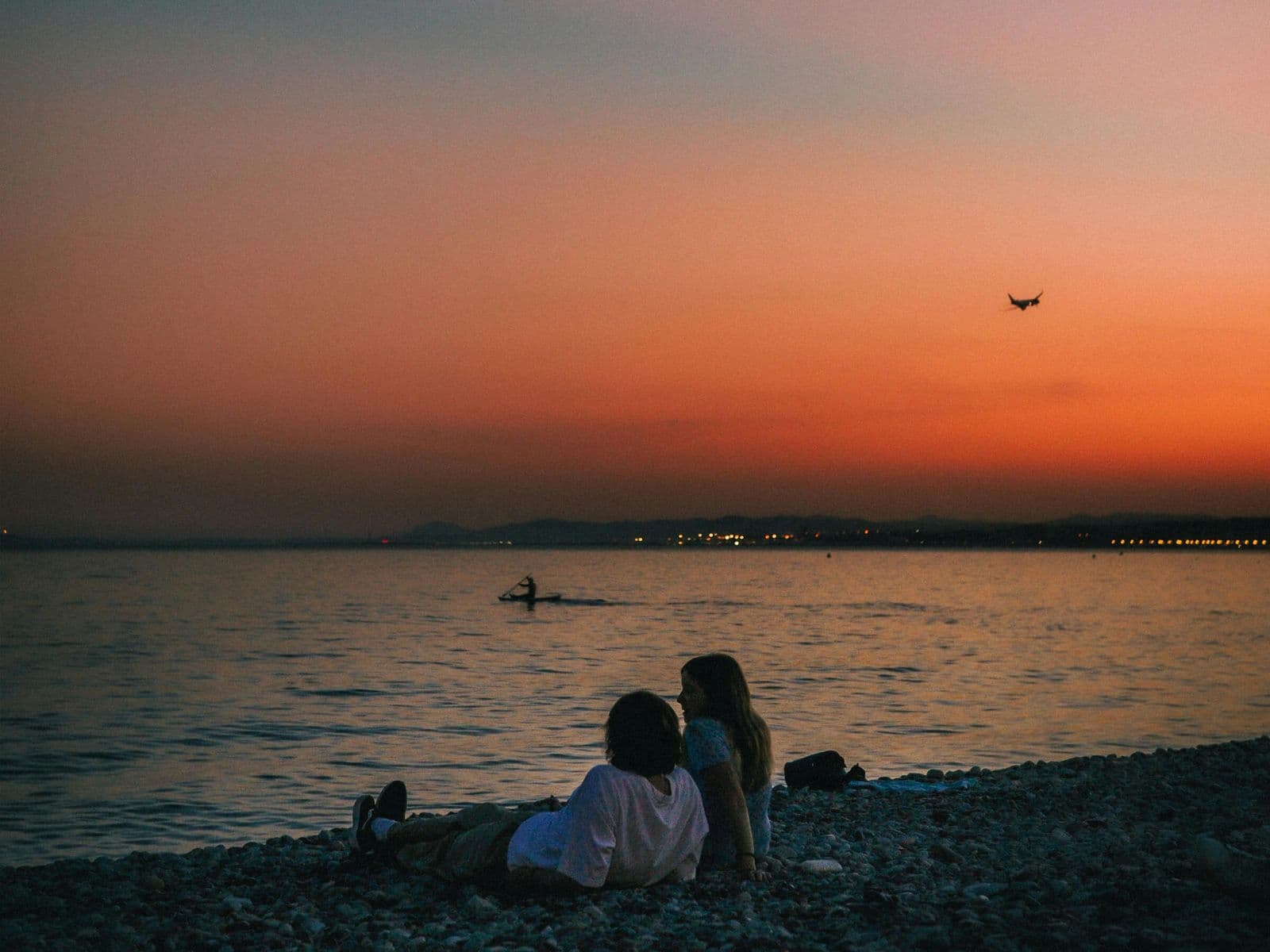 Harlowe – Sunset beach scene with two people sitting on a pebble shore watching a kayaker and a plane overhead; not clearly related to the building itself.