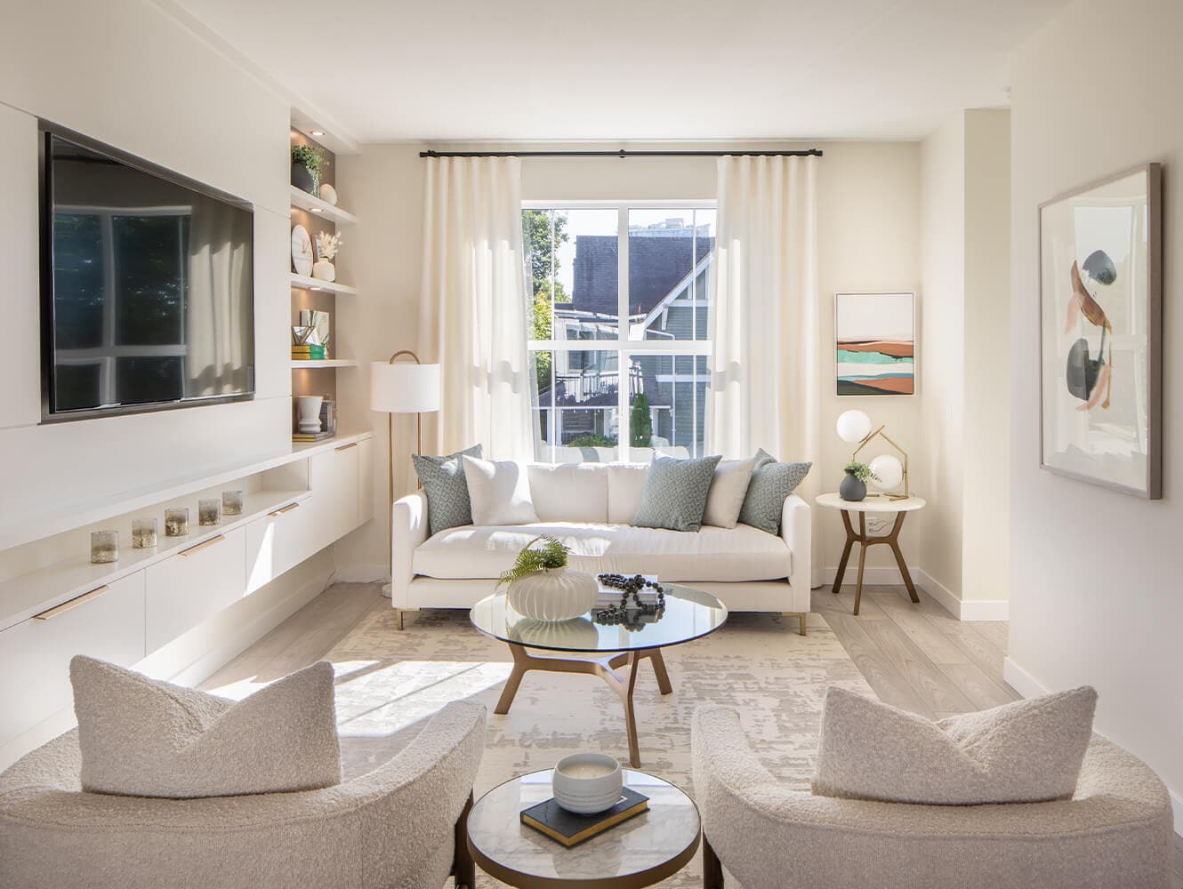 Portside – A bright, sunlit living room featuring a white sofa with textured blue throw pillows, two bouclé accent chairs, and a glass-top coffee table centered on an area rug. Built-in white cabinetry with floating shelves and a wall-mounted flat-screen television line one side, while a window framed by sheer white drapes fills the space with natural daylight.