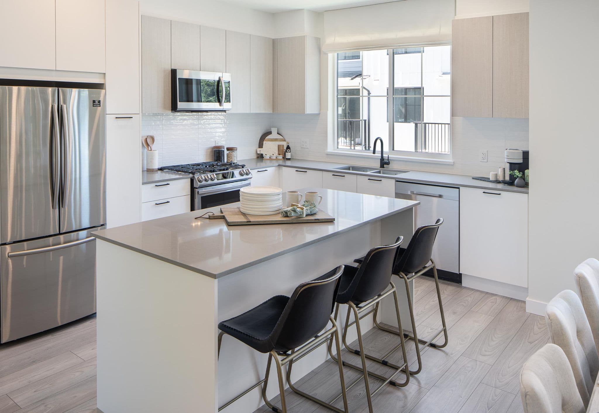 Portside – A modern, open-concept kitchen featuring light wood upper cabinets, white lower cabinets, and grey quartz countertops around a central kitchen island with three black bar stools. Stainless steel appliances, light laminate flooring, and natural daylight streaming in from a large window create a bright and clean aesthetic.