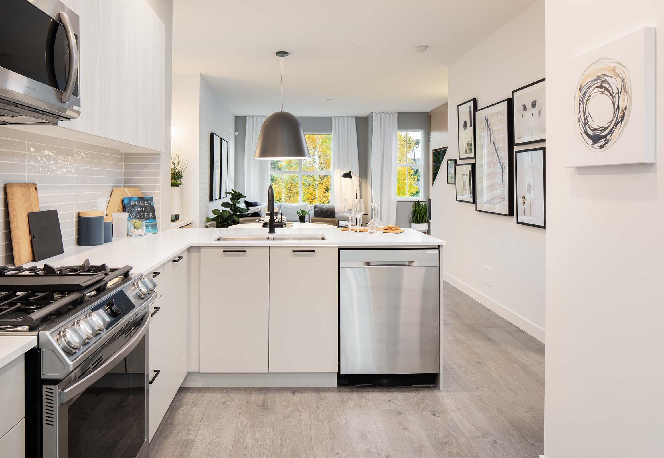 Portside – A modern open-concept kitchen featuring light oak-textured lower cabinetry, white quartz countertops, stainless steel appliances, and a black matte faucet under a matte dome pendant light. The view extends past the kitchen island into a bright living room with framed wall art, light wood-look flooring, and windows framed by white curtains showing autumn foliage outside.