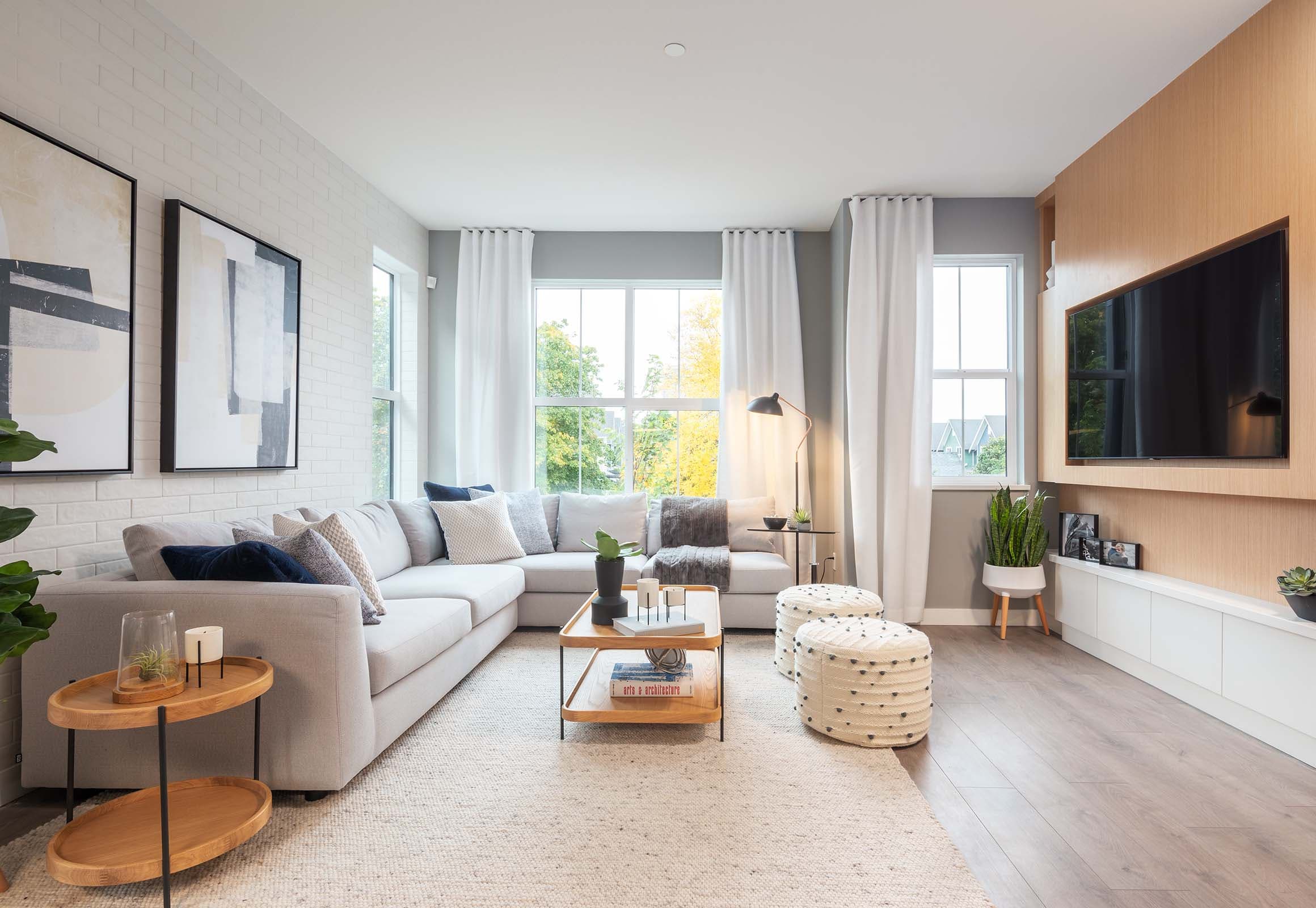 Portside – A modern, sunlit living room featuring a large light gray sectional sofa, a textured area rug, and a light wood coffee table paired with woven ottomans. The space includes a white painted brick accent wall with framed abstract art, a wall-mounted TV framed in light wood paneling, and large windows with floor-to-ceiling white drapes revealing autumn trees outside.