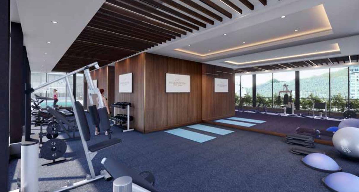 Millennium Central Lonsdale Condos – A fitness centre with weight machines, dumbbells, yoga mats, and floor-to-ceiling windows overlooking mountains and water.
