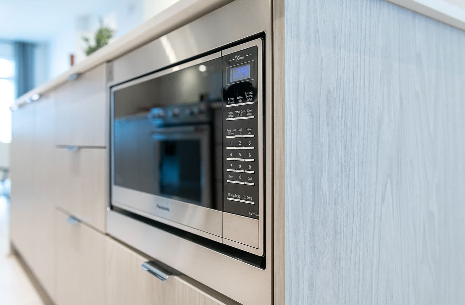 Chelsea at The Boroughs – Close-up of a built-in stainless-steel microwave in a kitchen island with light wood cabinetry.