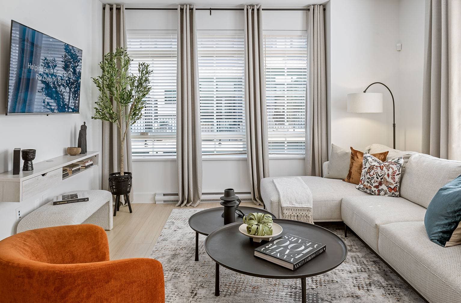 Chelsea at The Boroughs – Living room with a beige sectional sofa, orange accent chair, wall-mounted TV, and a bay window with blinds.