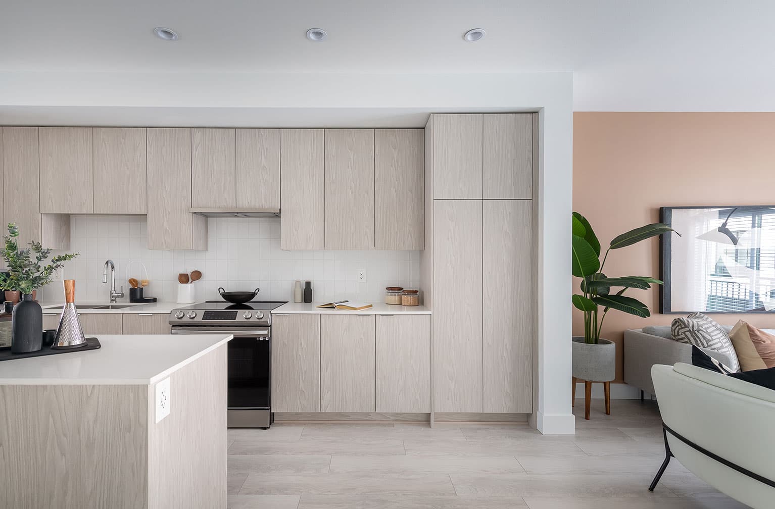Chelsea at The Boroughs – Kitchen with light wood cabinetry and white tile backsplash, opening to a living room with a peach-colored accent wall.