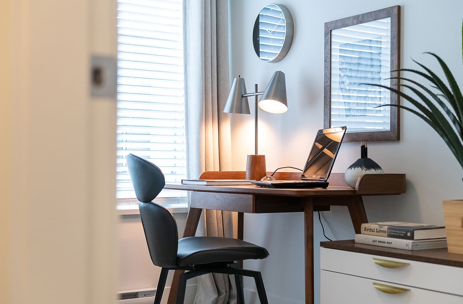 Chelsea at The Boroughs – Small home office nook with a wood desk, laptop, round mirror, and framed art near a window.