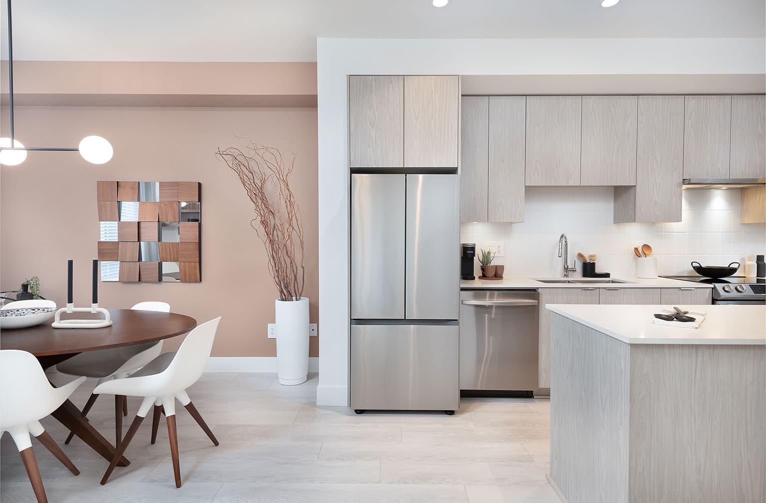 Chelsea at The Boroughs – Open kitchen with a stainless-steel fridge and dishwasher, next to a dining area with a peach accent wall and round table.