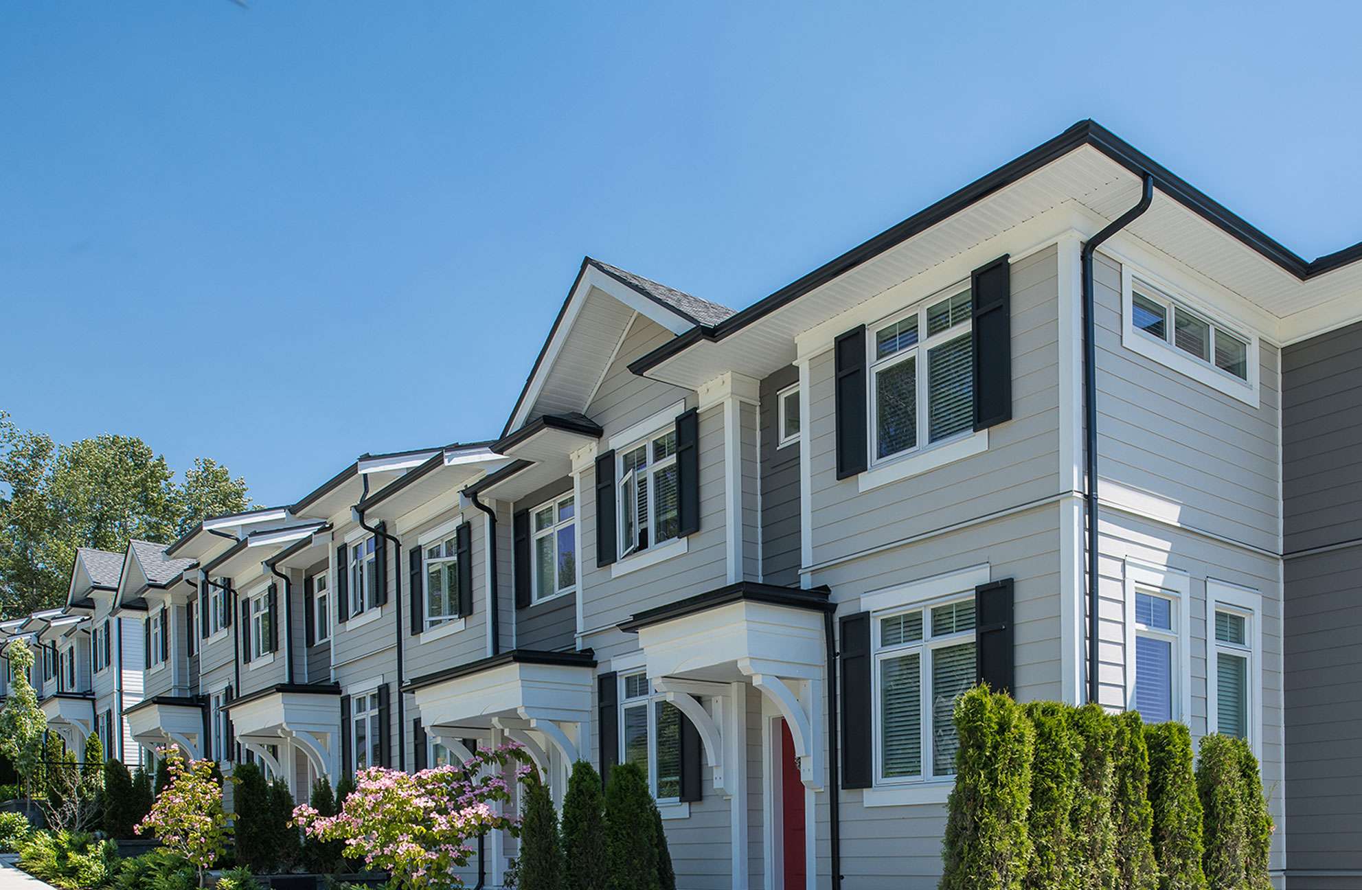 Chelsea at The Boroughs – Daytime view of a row of two-story townhomes with beige siding, dark shutters, and gabled roofs along a landscaped street.