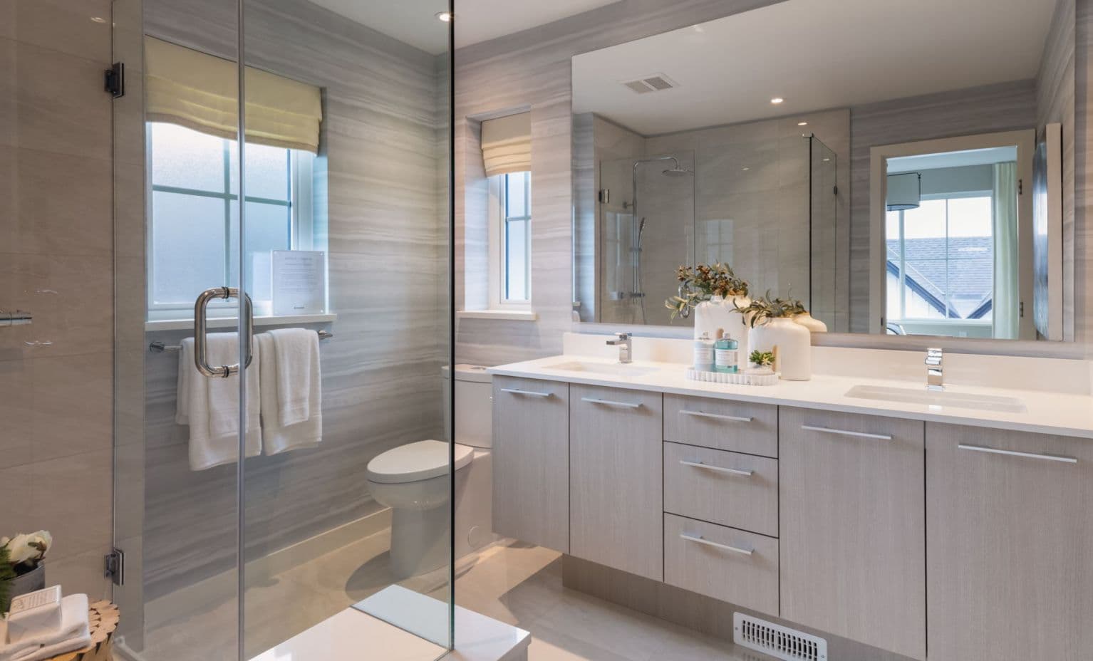 Ridgewood – Photo of a bathroom with a glass-enclosed shower, a white double vanity, and gray stone-look tile on the walls and floor.