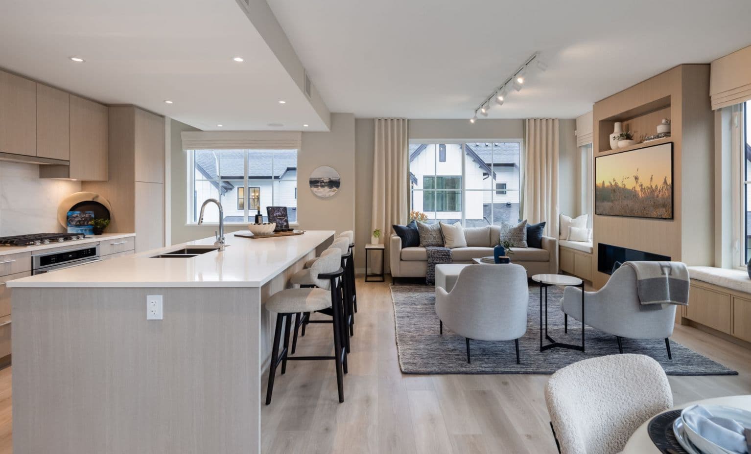 Ridgewood – Photo of an open living and kitchen area with a gray sectional sofa, wall-mounted TV, a window seat, and neighboring houses visible through the windows.