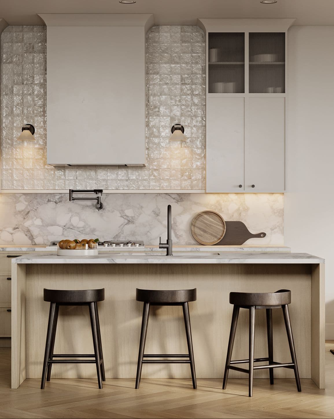 Edwyn – A contemporary kitchen design featuring a light wood island with three dark wooden barstools and a white marble countertop. In the background, a white marble backsplash is paired with glossy textured square tiles, warm sconce lighting, and pale minimalist cabinetry.