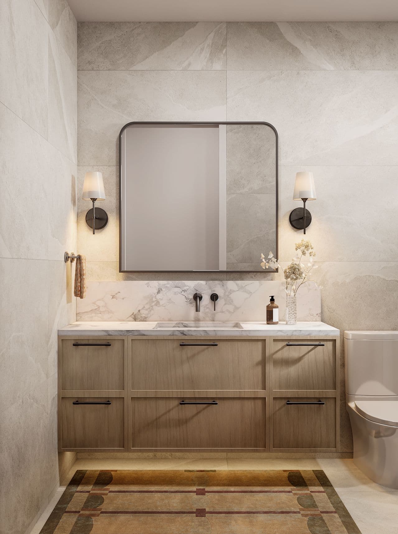 Edwyn – A contemporary bathroom vanity area featuring light wood floating cabinetry with black hardware, a white marble countertop with an integrated sink, and wall-mounted black fixtures. Large-format beige stone tiles cover the walls, complemented by a rounded rectangular mirror flanked by warm sconces and a patterned rug on the floor.