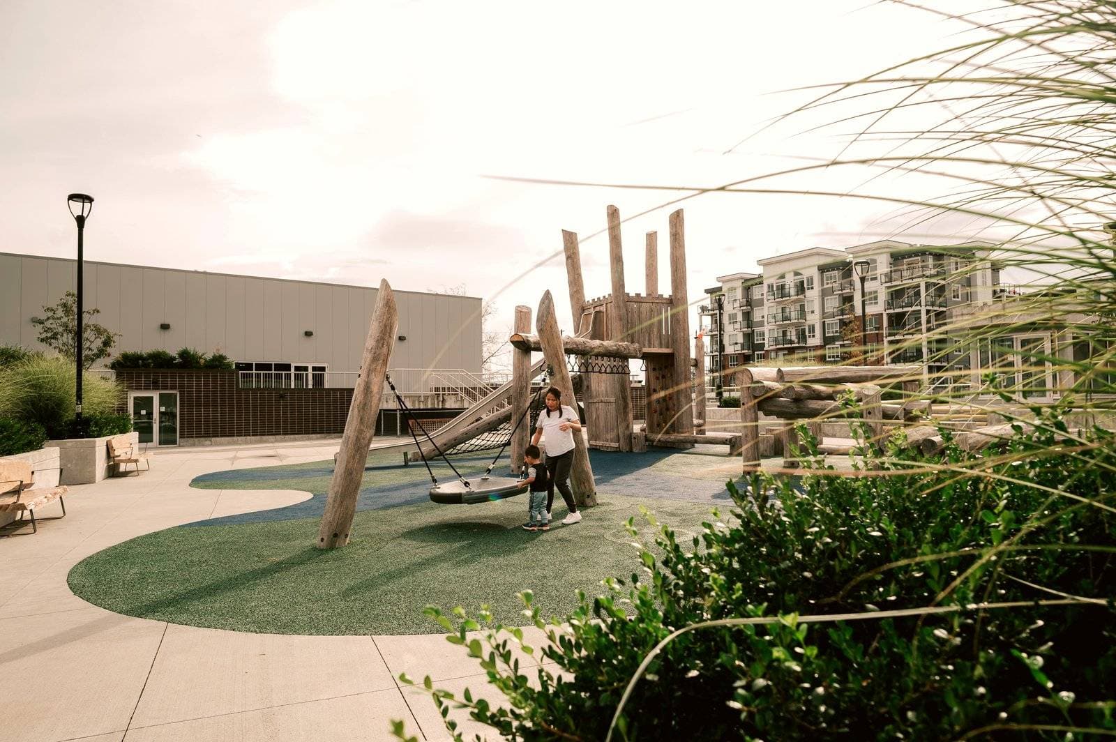Riverstone – An outdoor community playground featuring rustic log play structures, climbing ropes, a disc swing, and soft green safety surfacing set in an open courtyard. A woman and child interact by the swing, with modern residential apartment buildings and landscaped grasses visible in the background under soft daylight.