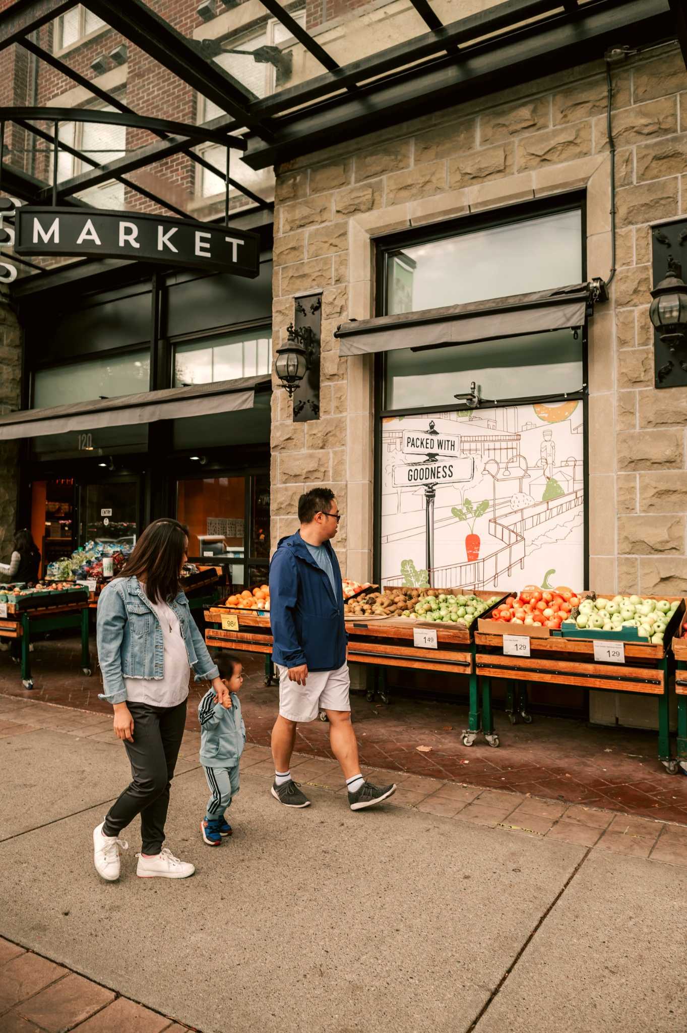 Riverstone – A family walks along a pedestrian sidewalk in front of a ground-level market featuring outdoor displays of fresh fruit. The building exterior features rustic stone masonry, black-framed storefront windows, a glass-and-steel canopy overhead, and charming lantern-style light fixtures.