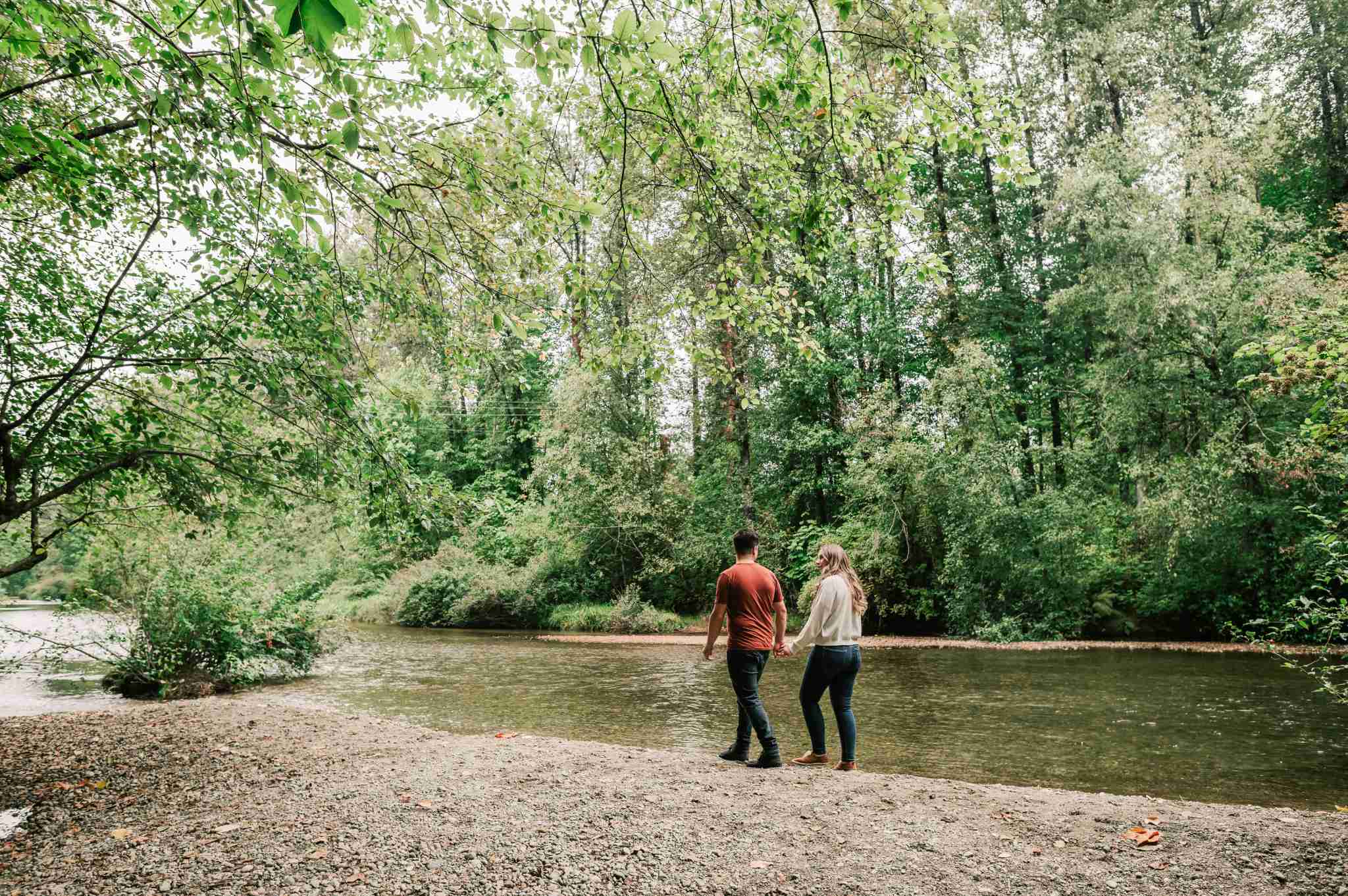 Riverstone – A couple walks hand-in-hand along a gravel riverbank beside a calm, clear flowing river. Lush green deciduous trees and dense forest canopy surround the tranquil outdoor natural setting in soft daylight.