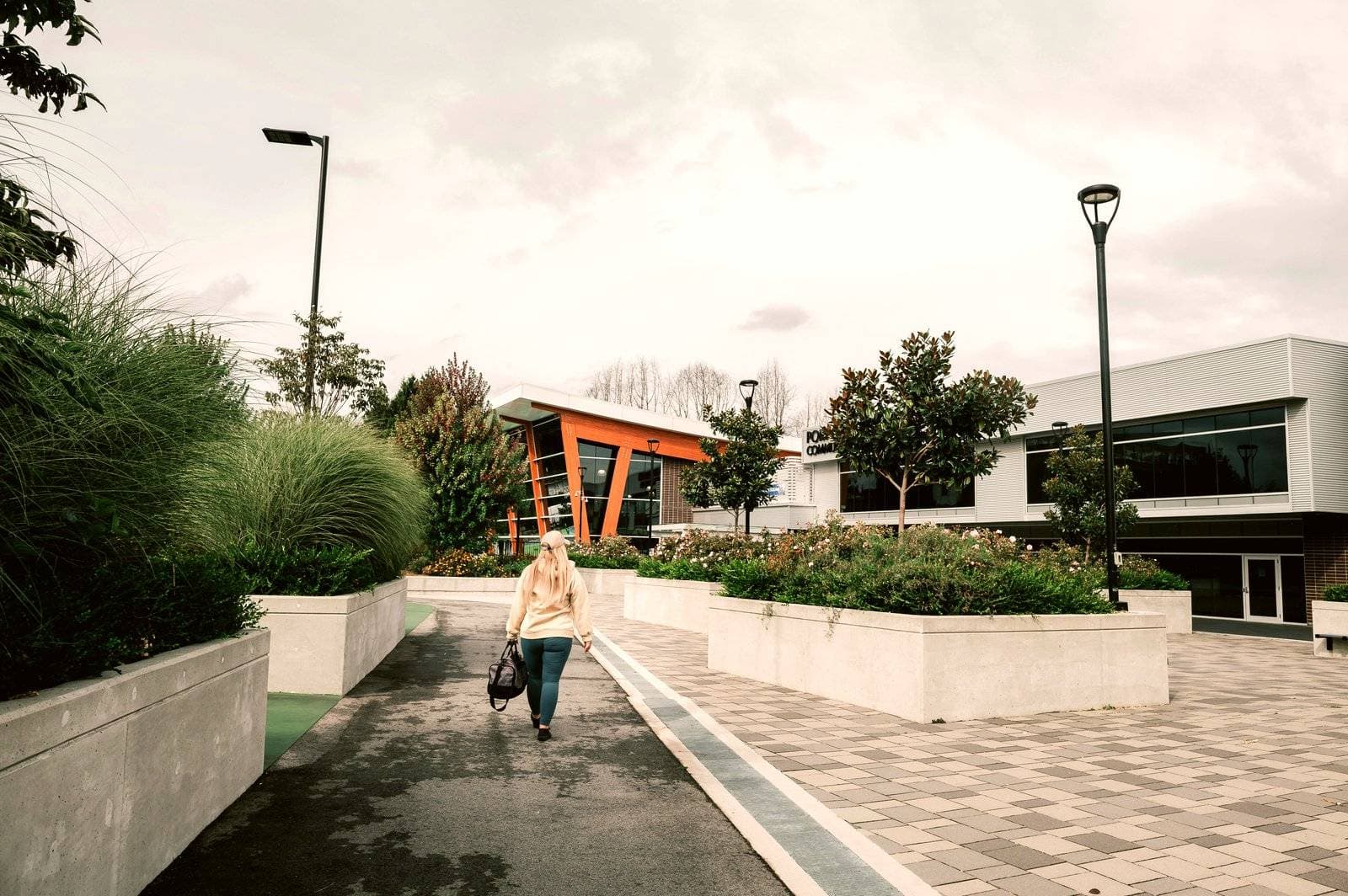 Riverstone – A pedestrian walks along a paved pathway lined with concrete planter boxes, manicured shrubs, and ornamental grasses. In the background, modern commercial or community buildings with large glass windows and orange architectural accents sit under an overcast sky.