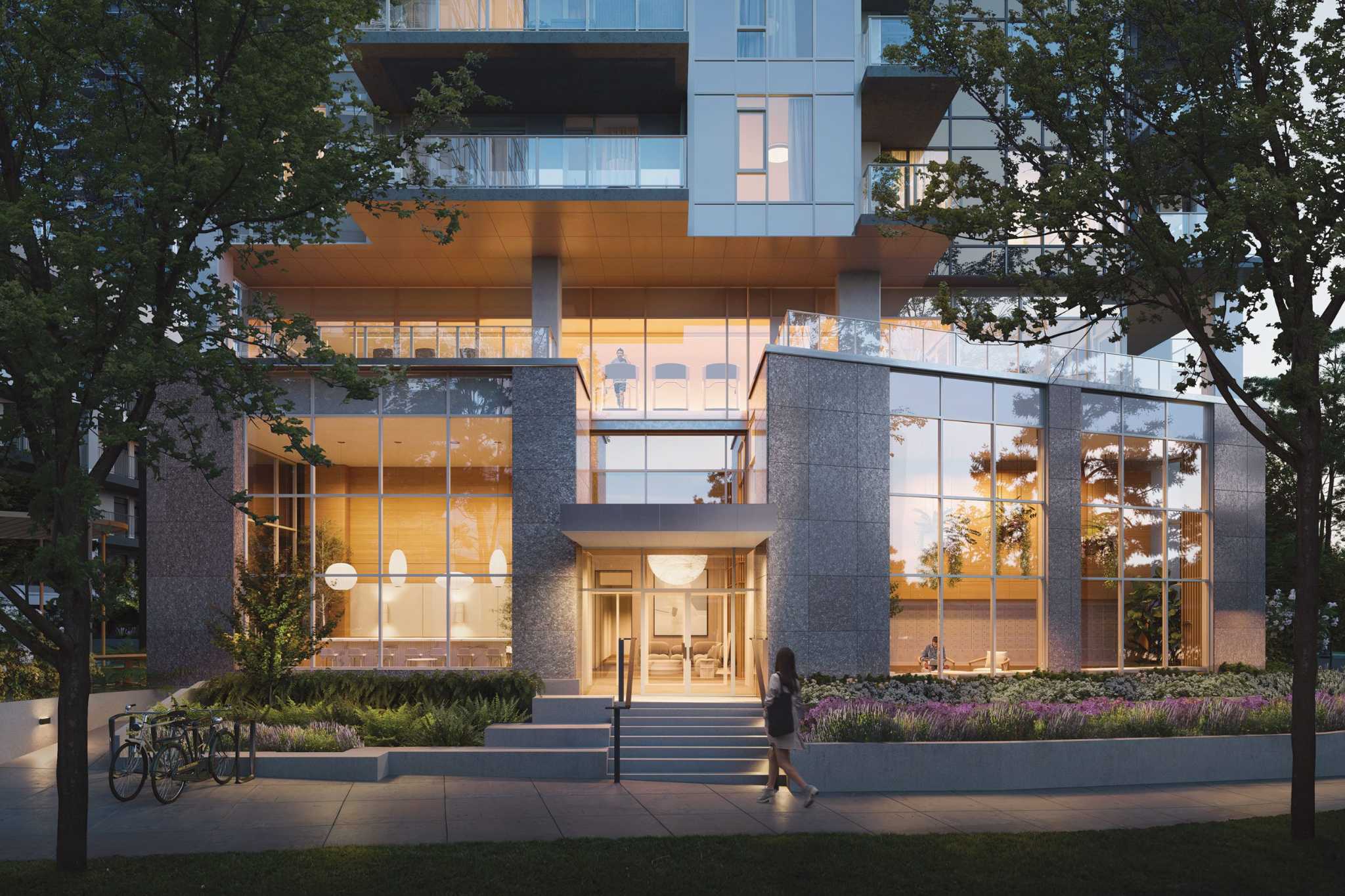 SOCO Two – A dusk rendering of the building's stone-clad entrance lobby with tall glass windows showing a lit lounge inside, bike racks, and a person walking up to the door.
