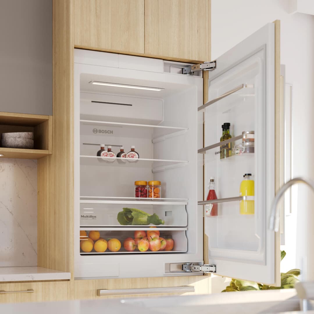 Astrid – A close-up view of a modern integrated Bosch refrigerator standing open within warm light-wood kitchen cabinetry. The brightly lit fridge interior is stocked with fresh produce and condiments, set beside a white quartz countertop and marble tile backsplash.