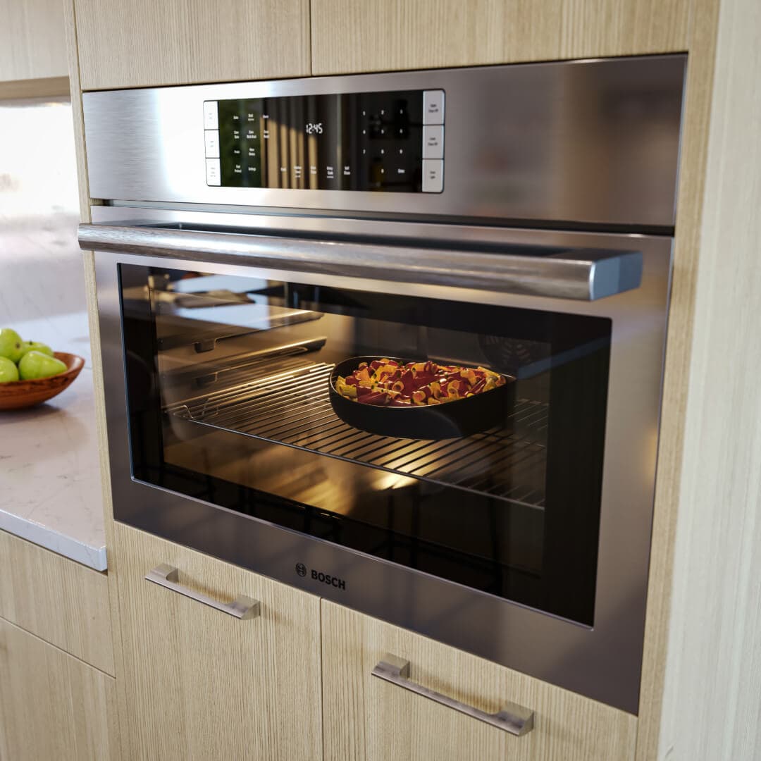 Astrid – A close-up shot features a modern, built-in stainless steel Bosch wall oven seamlessly integrated into light wood cabinetry. Inside the illuminated glass door, a dish bakes on a wire rack, while a bowl of green apples sits on the adjacent white marble countertop.