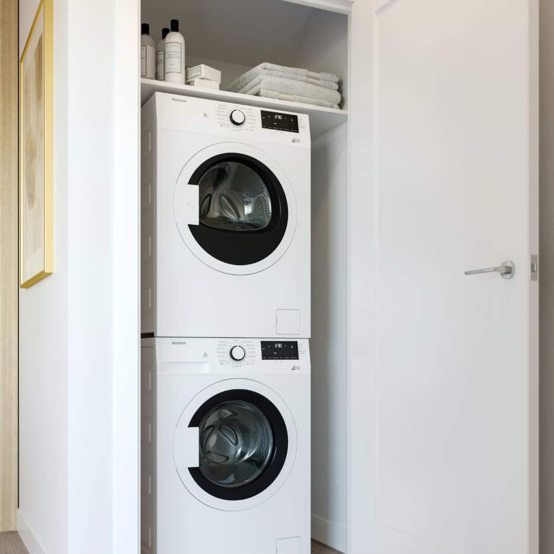 Astrid – A modern front-loading washer and dryer set stacked inside a clean white laundry closet with an open door. The upper shelf holds neatly folded towels and laundry supplies, creating a organized and functional residential space.