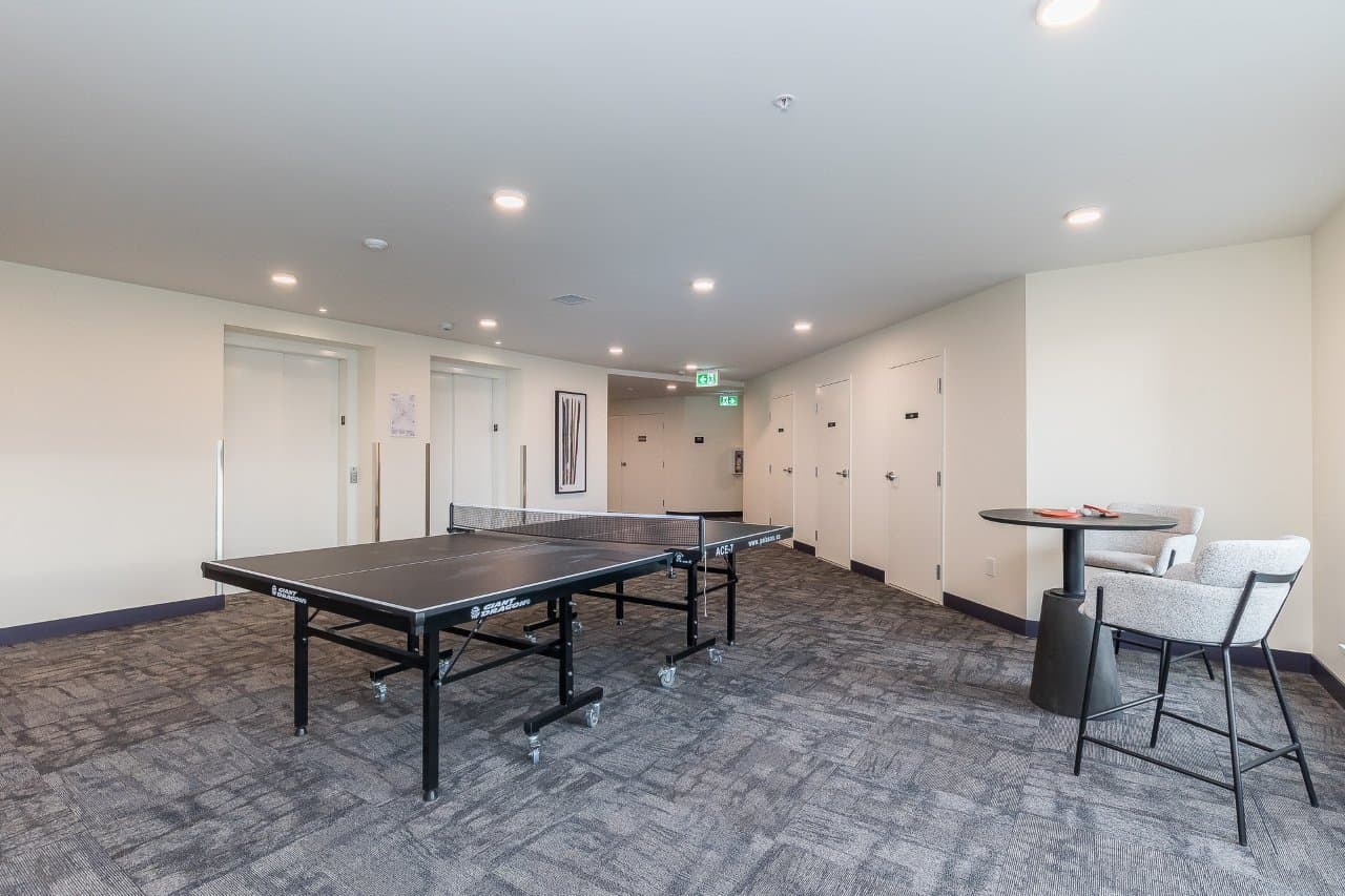 Mountainview Lane – Games lounge with a ping pong table, small seating area with armchairs, and a hallway with elevators in the background.