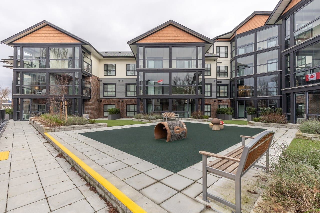 Mountainview Lane – Interior courtyard with a children's play area featuring a tunnel log climber and mushroom stools on green safety turf, surrounded by the building's glass-fronted units and a Canadian flag.