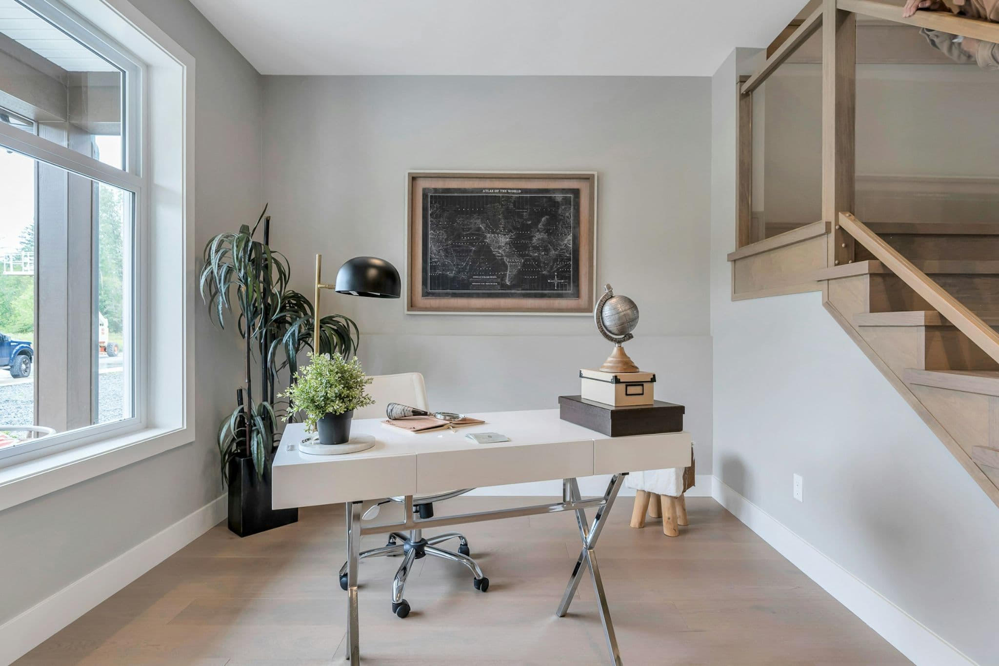 Emerald Ridge – Bright home office area with a white desk, chrome legs, large window, framed map artwork, and adjacent wooden staircase.