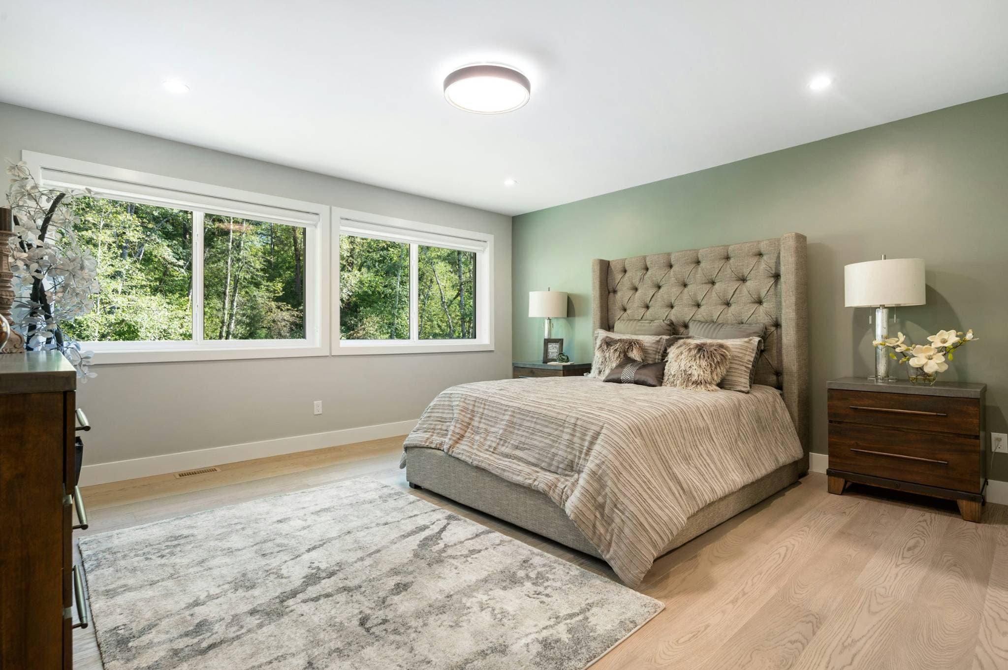 Emerald Ridge – Primary bedroom interior featuring a tufted upholstered bed, sage green accent wall, wooden nightstands, and large windows looking out to foliage.