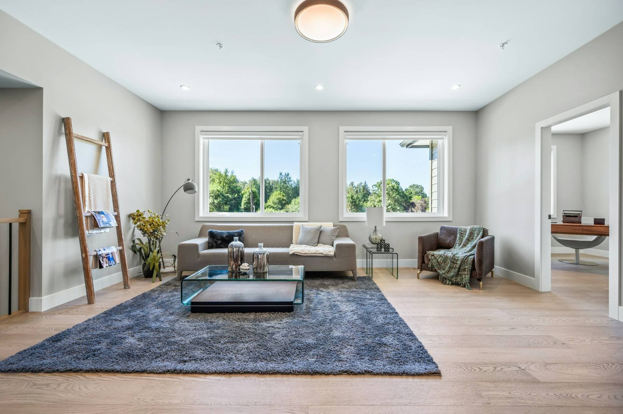 Emerald Ridge – Upper level seating area or secondary family room with a grey sofa, glass coffee table, plush area rug, and windows overlooking greenery.