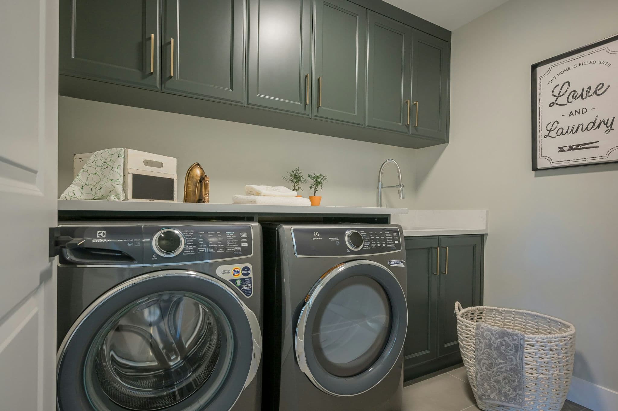 Emerald Ridge – Modern laundry room setup featuring dark green cabinetry, front-loading washer and dryer, laundry sink, and brass hardware.