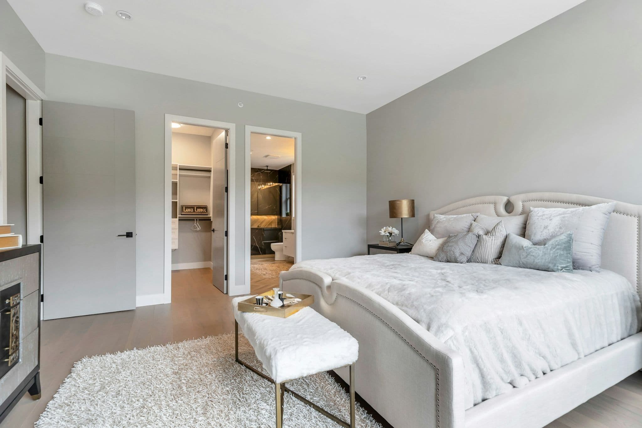 Emerald Ridge – Primary bedroom suite with upholstered headboard bed, plush bench, shag rug, and views into walk-in closet and ensuite bathroom.