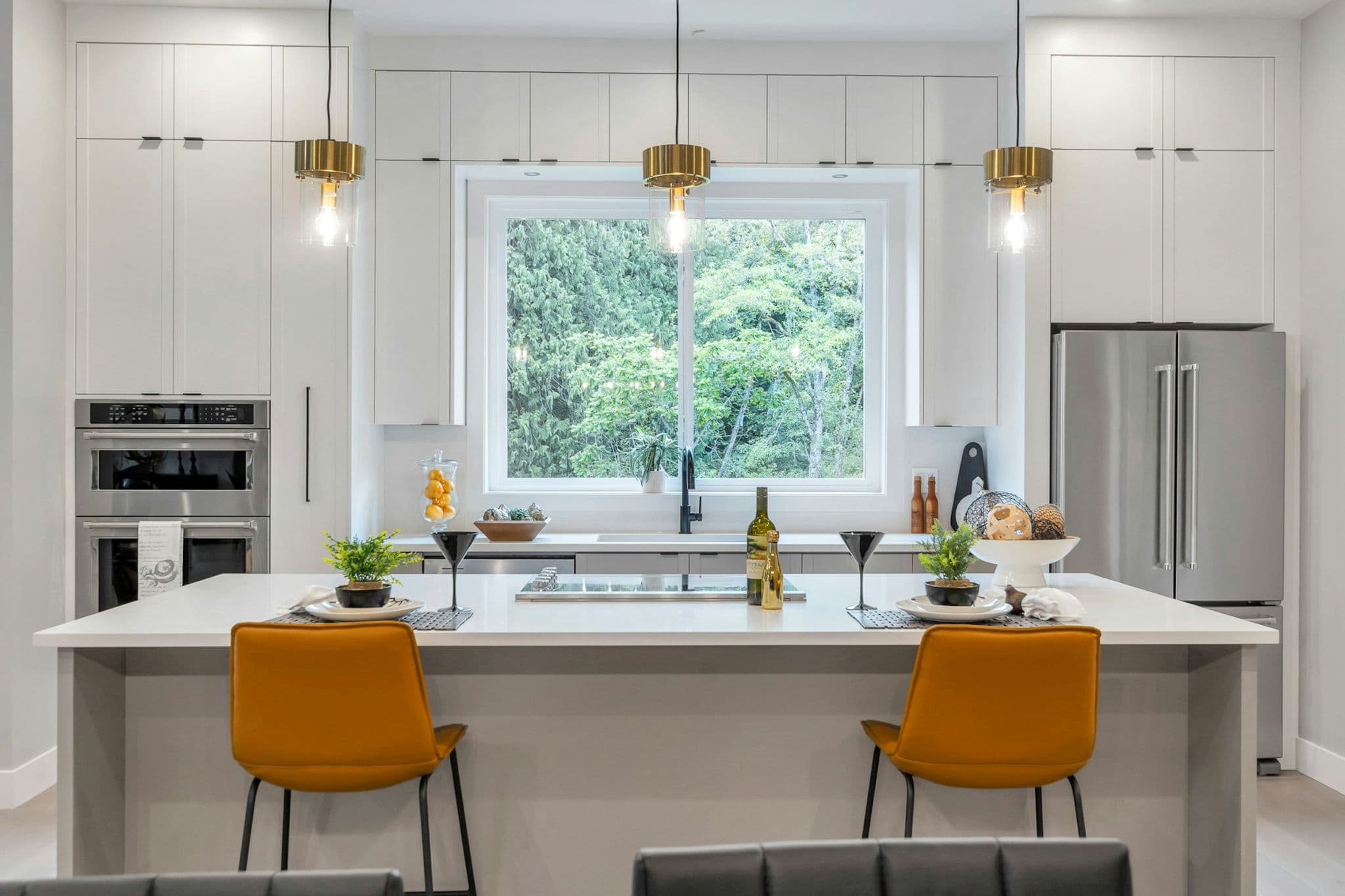 Emerald Ridge – Modern kitchen island featuring yellow bar stools, pendant light fixtures, white cabinetry, built-in double oven, and stainless steel refrigerator.