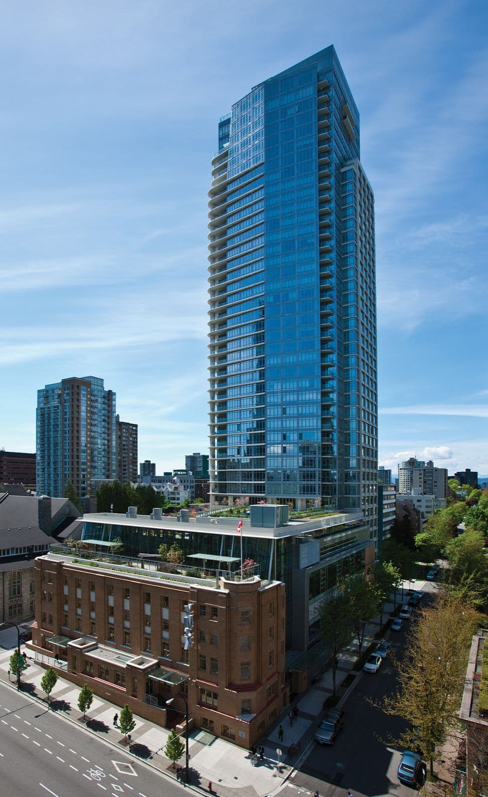 Tesoro – Elevated view of a tall blue glass residential tower rising behind a restored brick heritage building with a rooftop terrace, city skyline in the background.