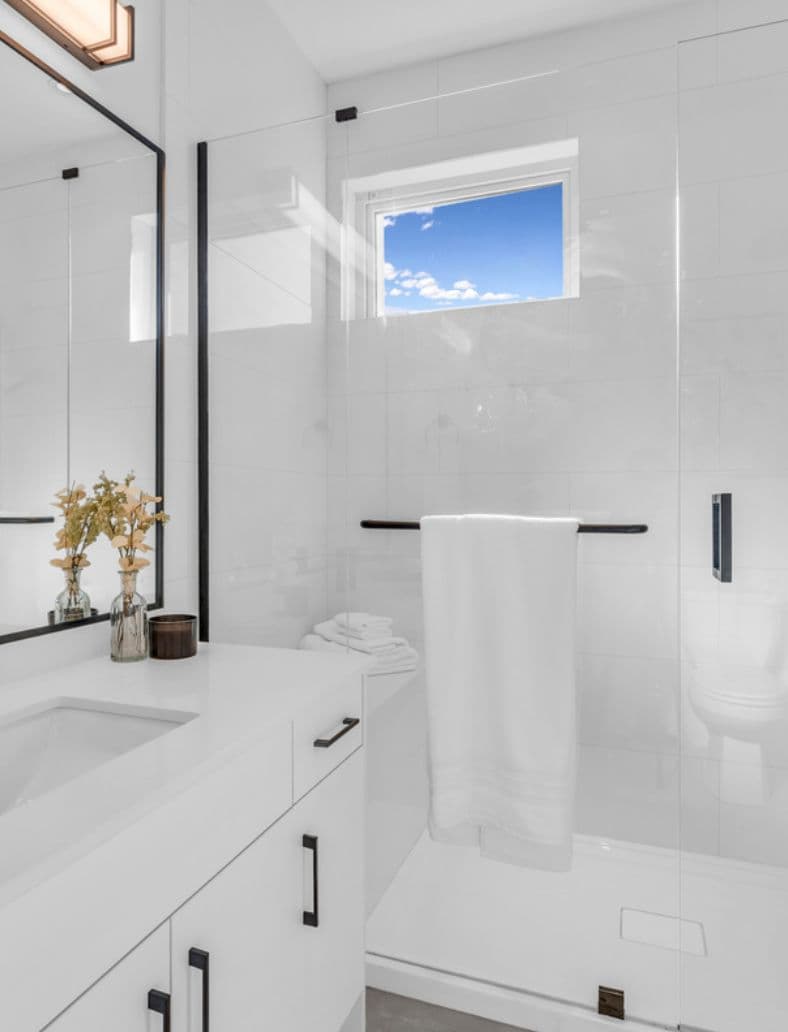 The Warwick – All-white bathroom with a glass-enclosed shower, hanging towels, a small window, and a vanity with black hardware in the foreground.
