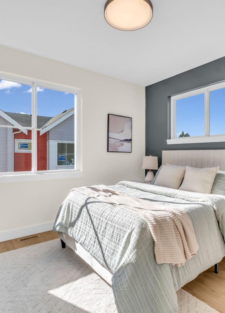 The Warwick – Bedroom with a dark accent wall behind an upholstered headboard, sage green bedding, a nightstand lamp, and two windows looking out at neighboring rooftops.